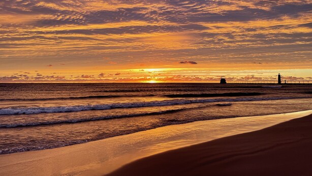 Una playa con un faro a lo lejos, olas suaves lamiendo la orilla y un atardecer con nubes dispersas.