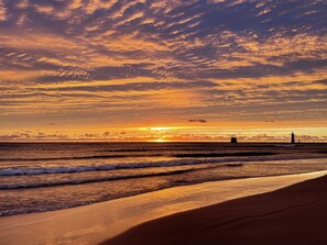 A beach with a lighthouse in the distance, waves gently lapping at the shore, and a sunset with scattered clouds. - Cozy home, minutes from lakeshore! (Spring Lake)