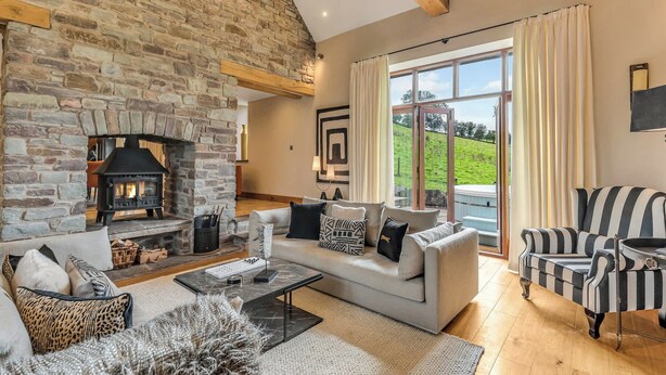 A cozy living room with a stone fireplace, a large window with curtains, a sofa, and a striped armchair. - Hilltop View with Hot Tub (Brecon)