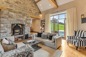 A cozy living room with a stone fireplace, a large window with curtains, a sofa, and a striped armchair. - Hilltop View with Hot Tub (Brecon)