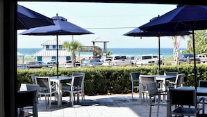 Un espace extérieur aménagé avec des tables et des chaises, des parasols, et une vue sur l’océan et les voitures garées.
