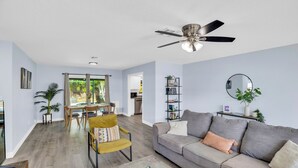 A modern living room with a grey sofa, a yellow armchair, a dining table with chairs, a ceiling fan, and a large window. - Relax and Unwind in 2 BR apt with Private Heated Pool (Sebastian)