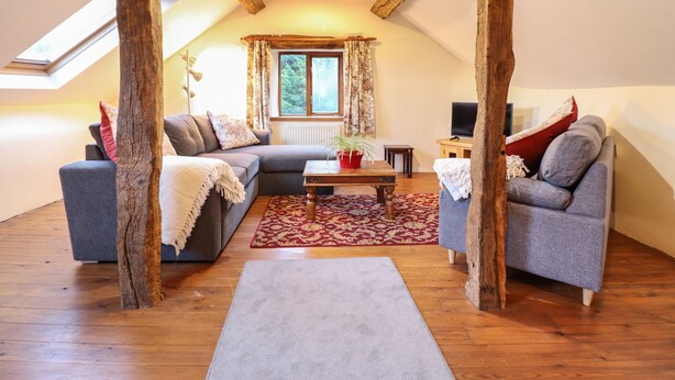 A cozy loft-style living room with a sloped ceiling, wooden beams, a large window, and two grey sofas with red cushions. - Yr Hen Felin (Oswestry)