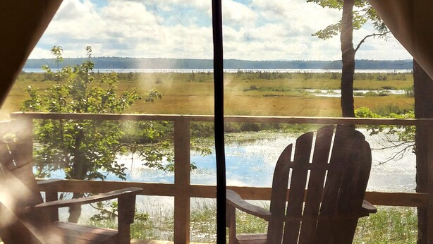 Eine Holzterrasse mit einem Stuhl, mit Blick auf einen See und ferne Bäume.