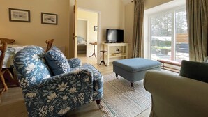 Una sala de estar con sofá, reposapiés y un televisor sobre un mueble bajo. Se ve un comedor con una mesa y sillas a través de una puerta abierta.