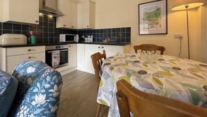 Una cocina compacta con alacenas blancas, un microondas y una estufa. Hay una mesa de comedor con mantel estampado de hojas y sillas de madera. Un cuadro enmarcado con un paisaje cuelga en la pared, y una lámpara de pie está a la derecha.