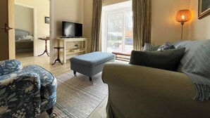 Una sala de estar con un sofá, un reposapiés y un televisor sobre un mueble bajo. Hay una puerta que da a un dormitorio y una ventana con cortinas.