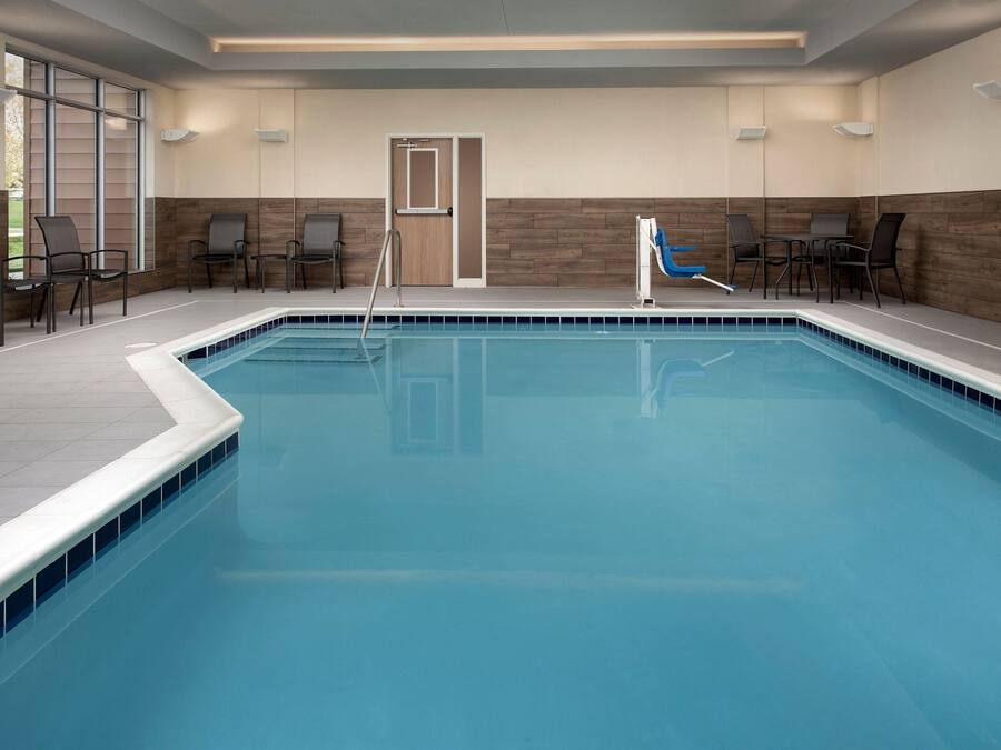 Una piscina interior con superficie de agua cristalina, rodeada por un solárium con baldosas. Se ven sillas y una puerta al fondo.