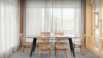 Un comedor con mesa de vidrio rodeada de sillas de madera, ventanas amplias con cortinas livianas y vistas al exterior.