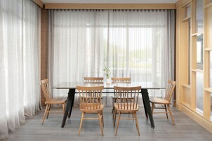 Un comedor con mesa de vidrio rodeada de sillas de madera, ventanas amplias con cortinas livianas y vistas al exterior.
