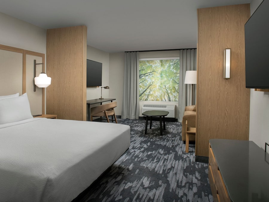A hotel room with a large bed, a desk, a chair, a TV, and a window with a view of trees.