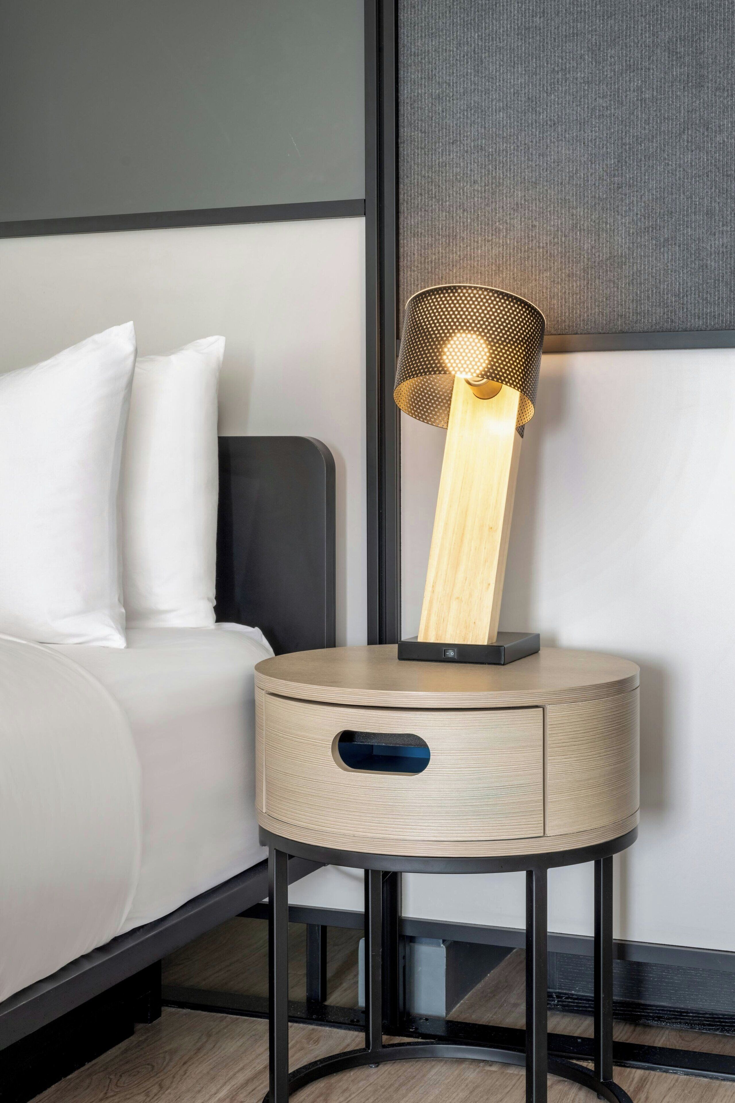 Una mesita de noche moderna con detalles de madera y una base de metal negro, junto a una cama blanca y un cabecero gris.