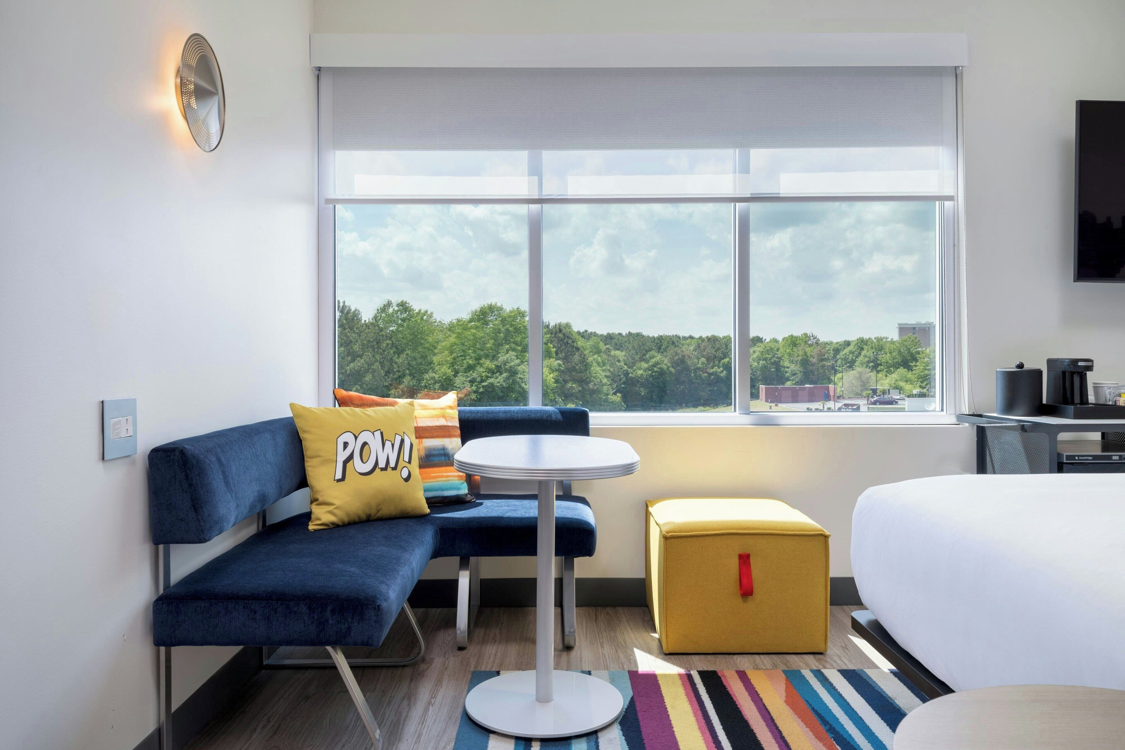 A modern hotel room with a sofa, a small round table, a yellow ottoman, and a bed.