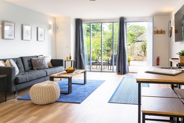 Un salon moderne comprenant un canapé, une table basse et un pouf, un coin repas avec une table et des chaises, et une grande porte-fenêtre coulissante donnant sur une terrasse extérieure.