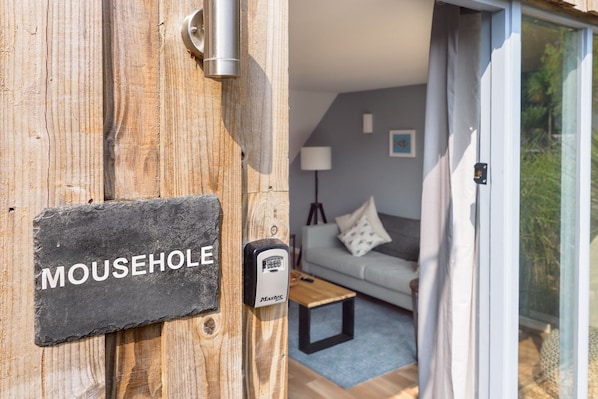 Une porte en bois portant l’inscription « MOUSEHOLE » et un intérieur moderne avec un canapé, une lampe et une petite table.