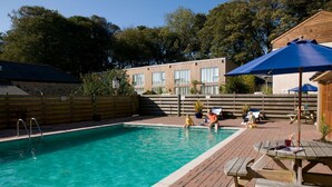 A swimming pool with a wooden deck, a picnic table, and a building with large windows in the background. - Charming Cornish cottage with private hot tub/shared pool, nr Falmouth/Helford (Falmouth)