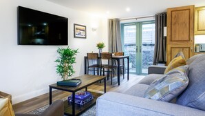 A living room with a sofa, a coffee table, a TV, a dining area with chairs, and a view through a glass door. - Charming Cornish cottage with private hot tub/shared pool, nr Falmouth/Helford (Falmouth)