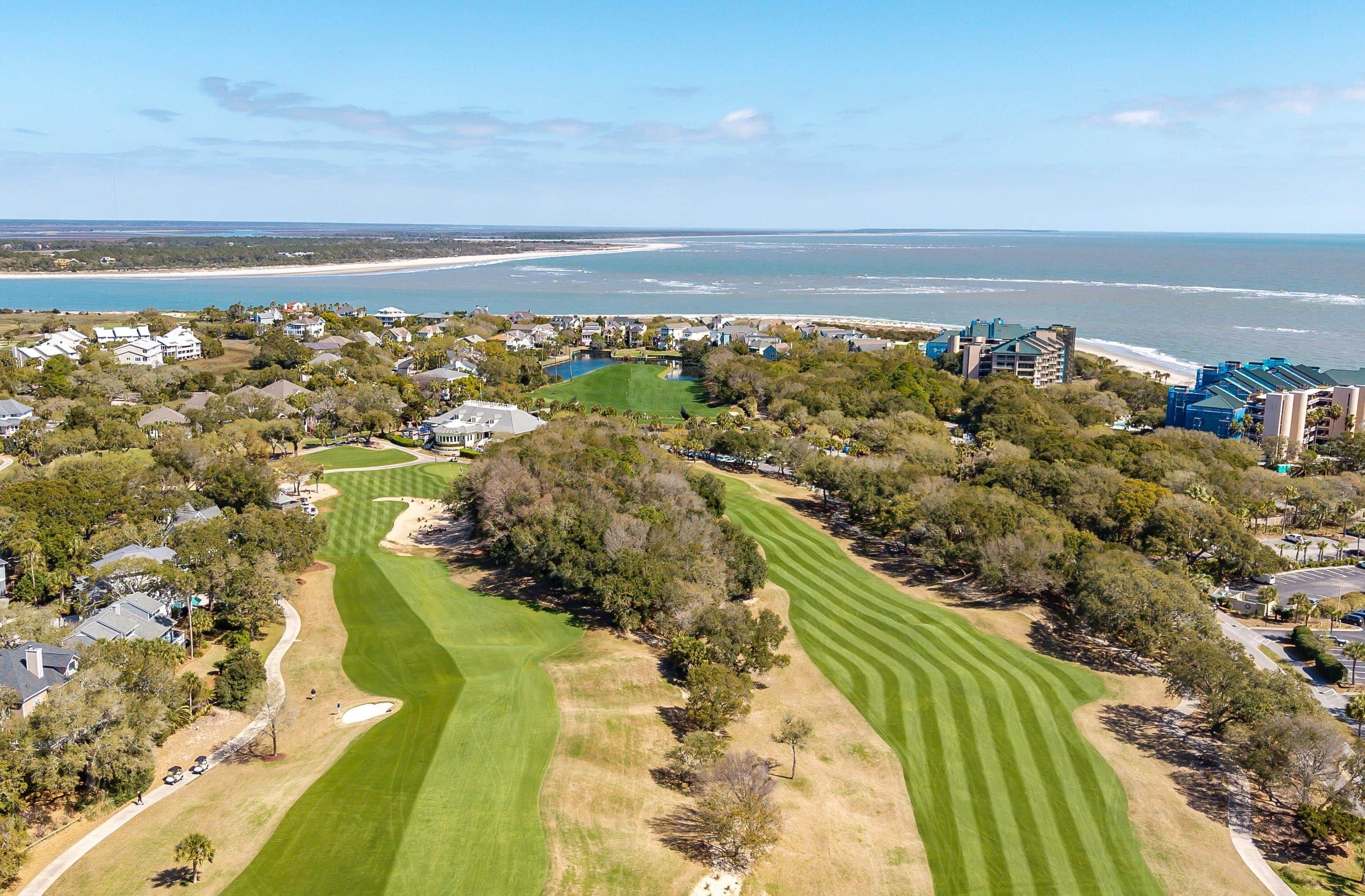Vue aérienne d’un terrain de golf bordé d’une plage, avec des immeubles résidentiels et un ciel dégagé.