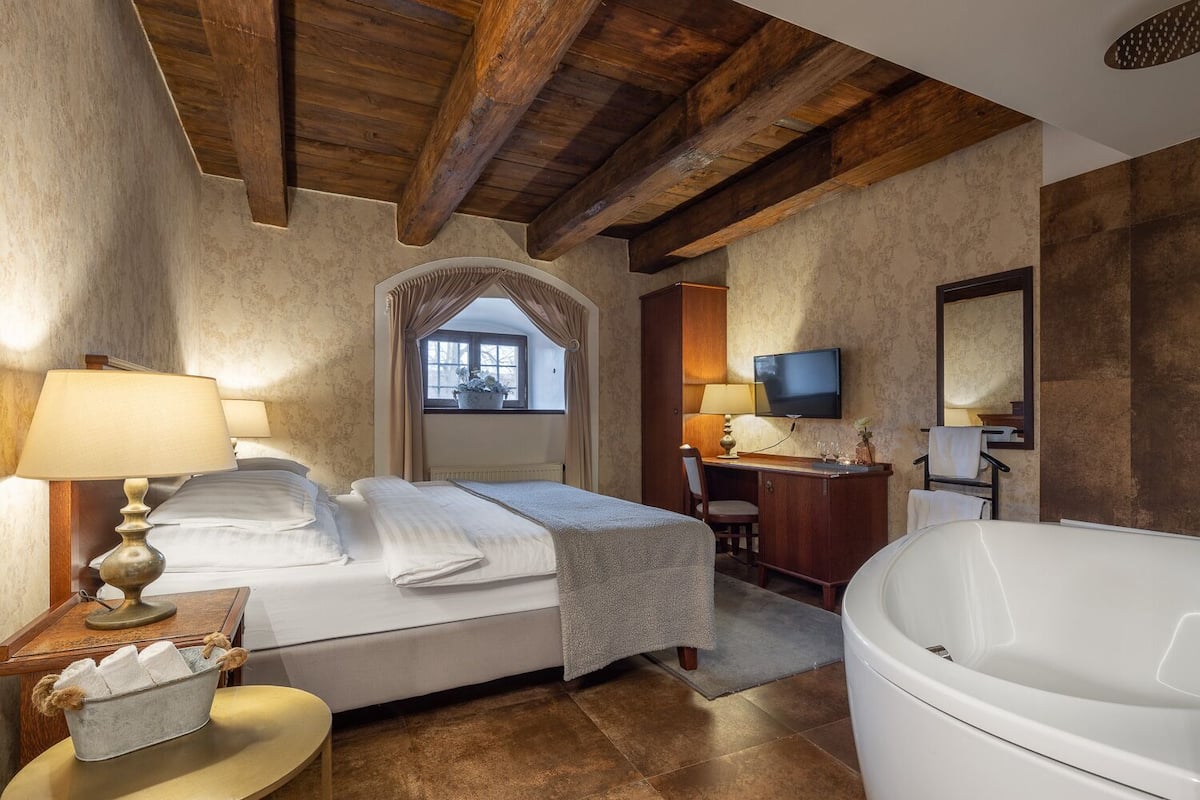 Une chambre d’hôtel avec un grand lit, un bureau avec une chaise, une télévision, une grande baignoire et une fenêtre avec des rideaux.