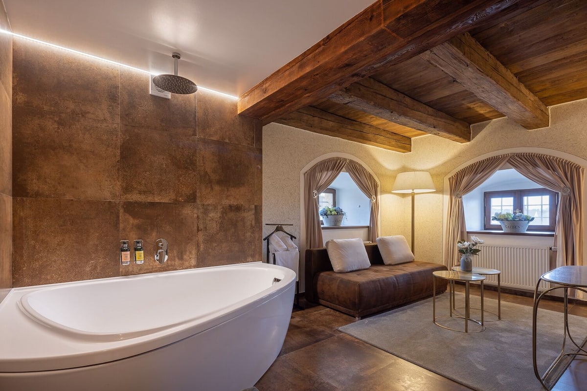 Une salle de bain spacieuse avec une grande baignoire, des poutres en bois au plafond et un coin salon confortable donnant sur un jardin, visible à travers une fenêtre.