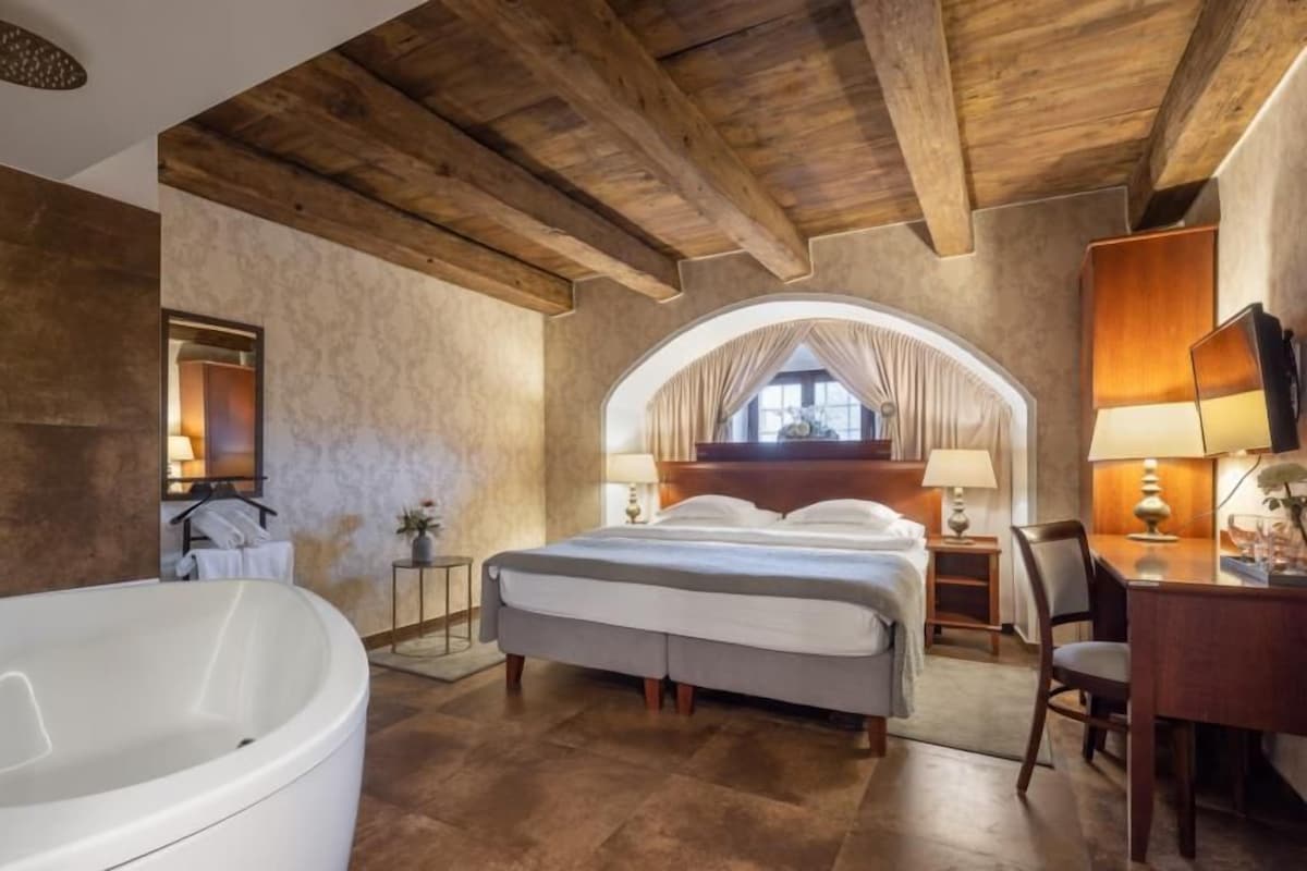 Un dormitorio con una cama grande, una zona de estar con escritorio y silla, una bañera y un amplio ventanal de arco.