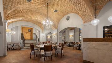 A spacious dining area with high ceilings and exposed brick walls, featuring a central chandelier and multiple tables set for dining.