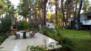 A patio area with a stone table and two benches, surrounded by lush greenery and tall trees.