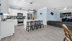 Eine moderne Küche mit weißen Schränken, einer zentralen Kochinsel mit Barhocker und einem Essbereich mit Sofa und Couchtisch.