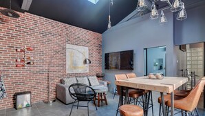 Ein modernes Interieur mit einer Ziegelwand, einem Essbereich mit Holztisch und Ledersesseln, einer Couch mit Kissen und einer Stehlampe.