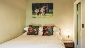A bedroom with a large bed, two floral pillows, a nightstand with a lamp, and a painting of cows on the wall.