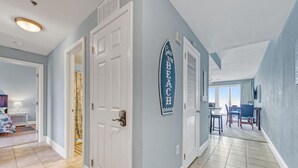 Un couloir avec une porte donnant sur une chambre, un coin repas avec des chaises, et une pancarte sur le mur indiquant : « Je suis à la plage. »