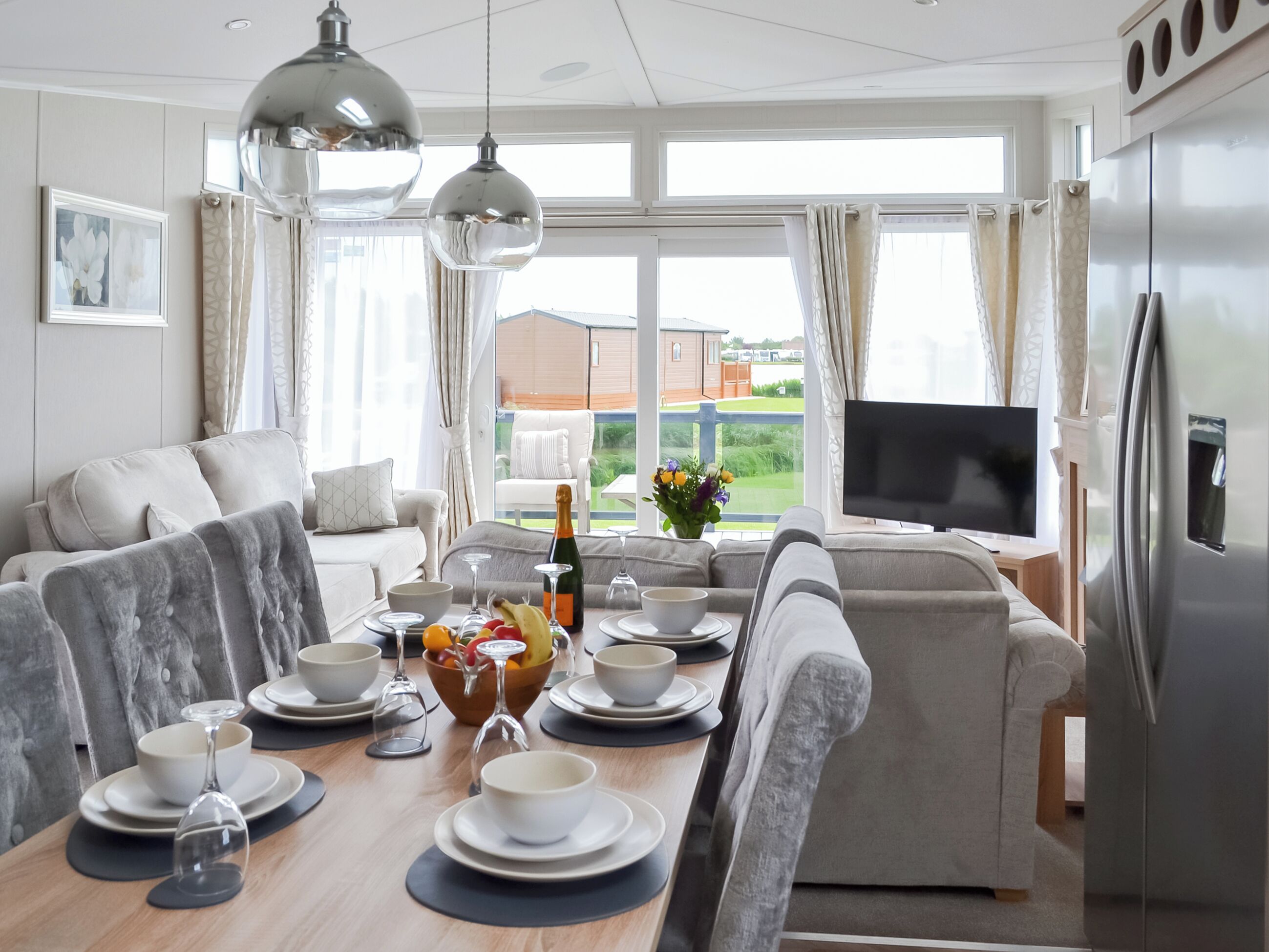 A modern living room with a dining area, a large window with curtains, a flat-screen TV, and a refrigerator.