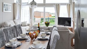 A modern living room with a dining area, a large window with curtains, a flat-screen TV, and a refrigerator. - Reel Paradise - UK37723 (Addlethorpe, near Skegness)