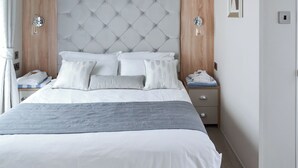 A neatly made bed with a tufted headboard, a bedside table with a lamp, and a nightstand with folded towels. - Reel Paradise - UK37723 (Addlethorpe, near Skegness)