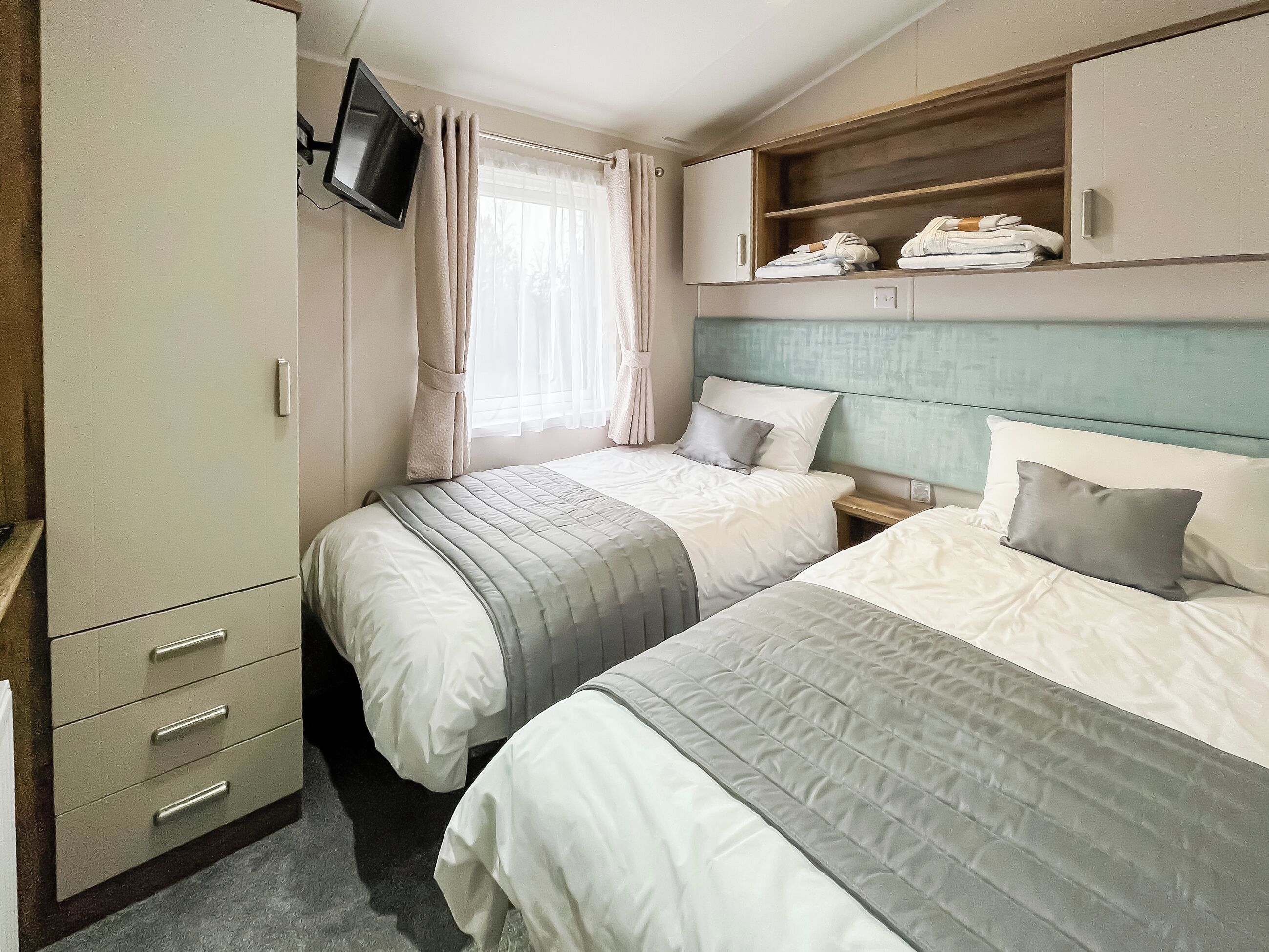 A bedroom with two beds, a TV mounted on the wall, and a wardrobe with drawers.