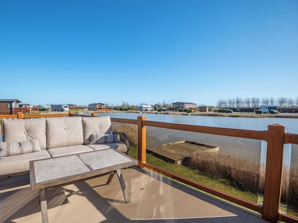 A deck with a sofa and table, overlooking a body of water with houses in the background.