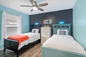 Quarto com duas camas, um ventilador de teto, janela com persianas e uma parede com luminária neon.