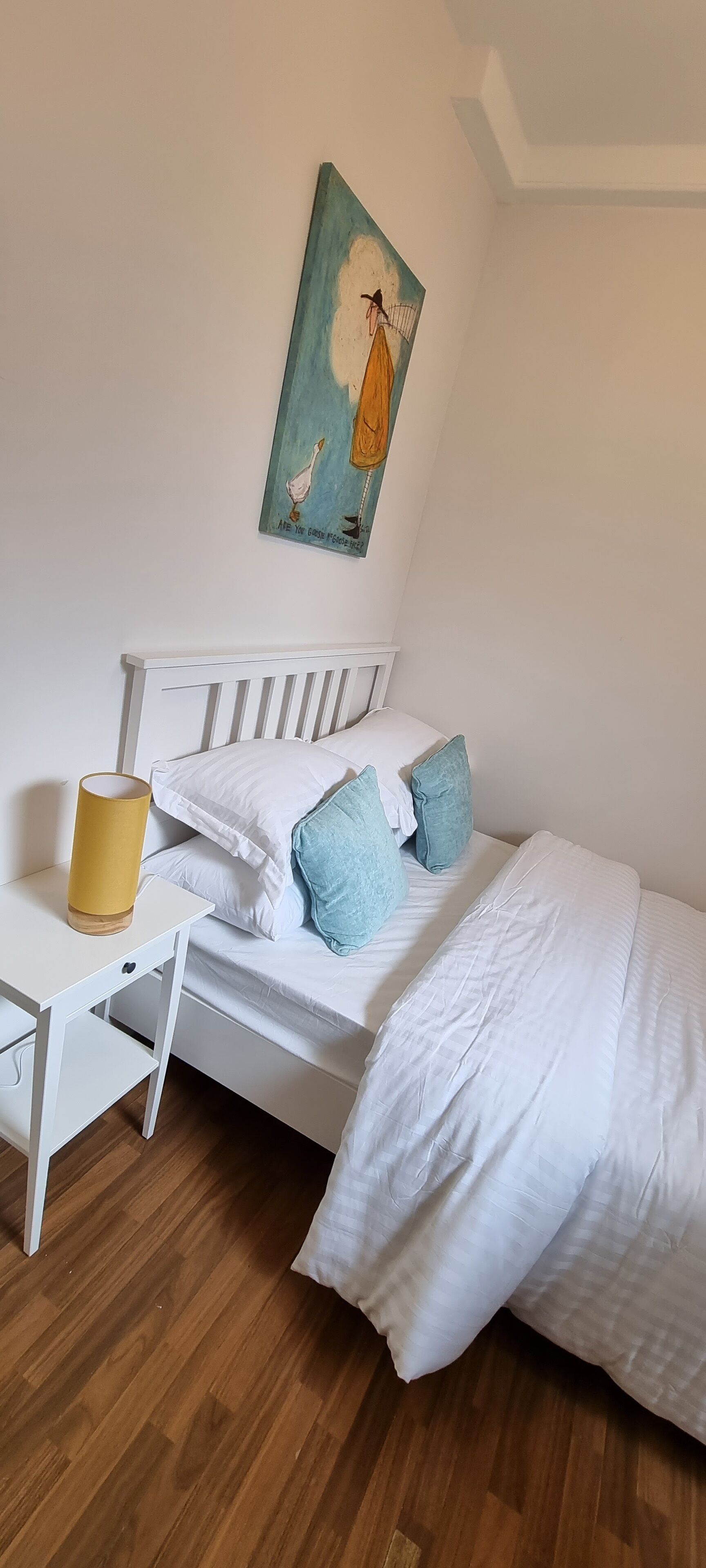 A neatly arranged bedroom with a white bed, a small bedside table with a lamp, and a painting on the wall.
