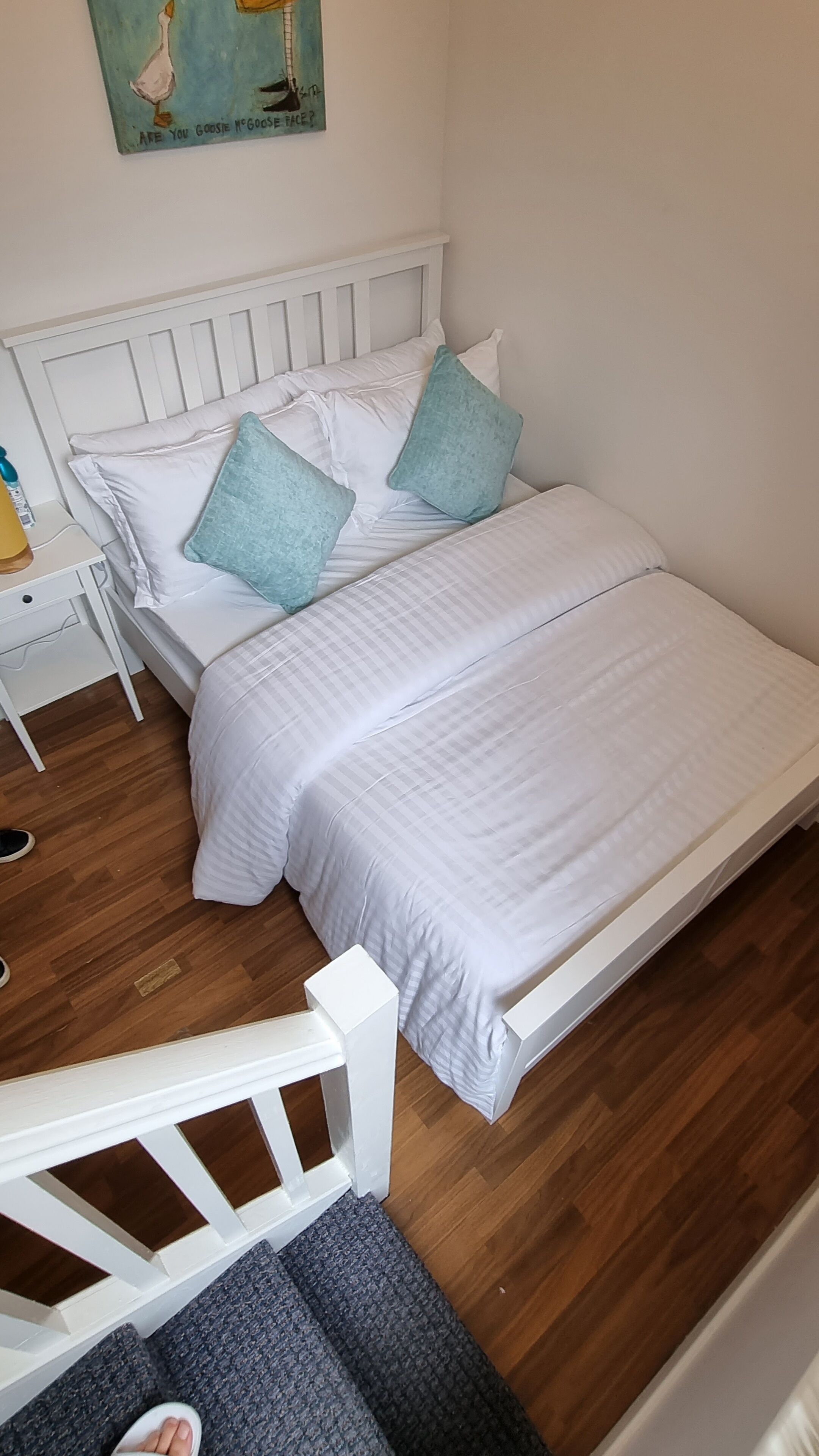 Ein ordentlich bezogenes Bett mit weißer Bettwäsche und zwei türkisfarbenen Kissen, ein weißer Nachttisch mit Lampe sowie ein Holzboden.