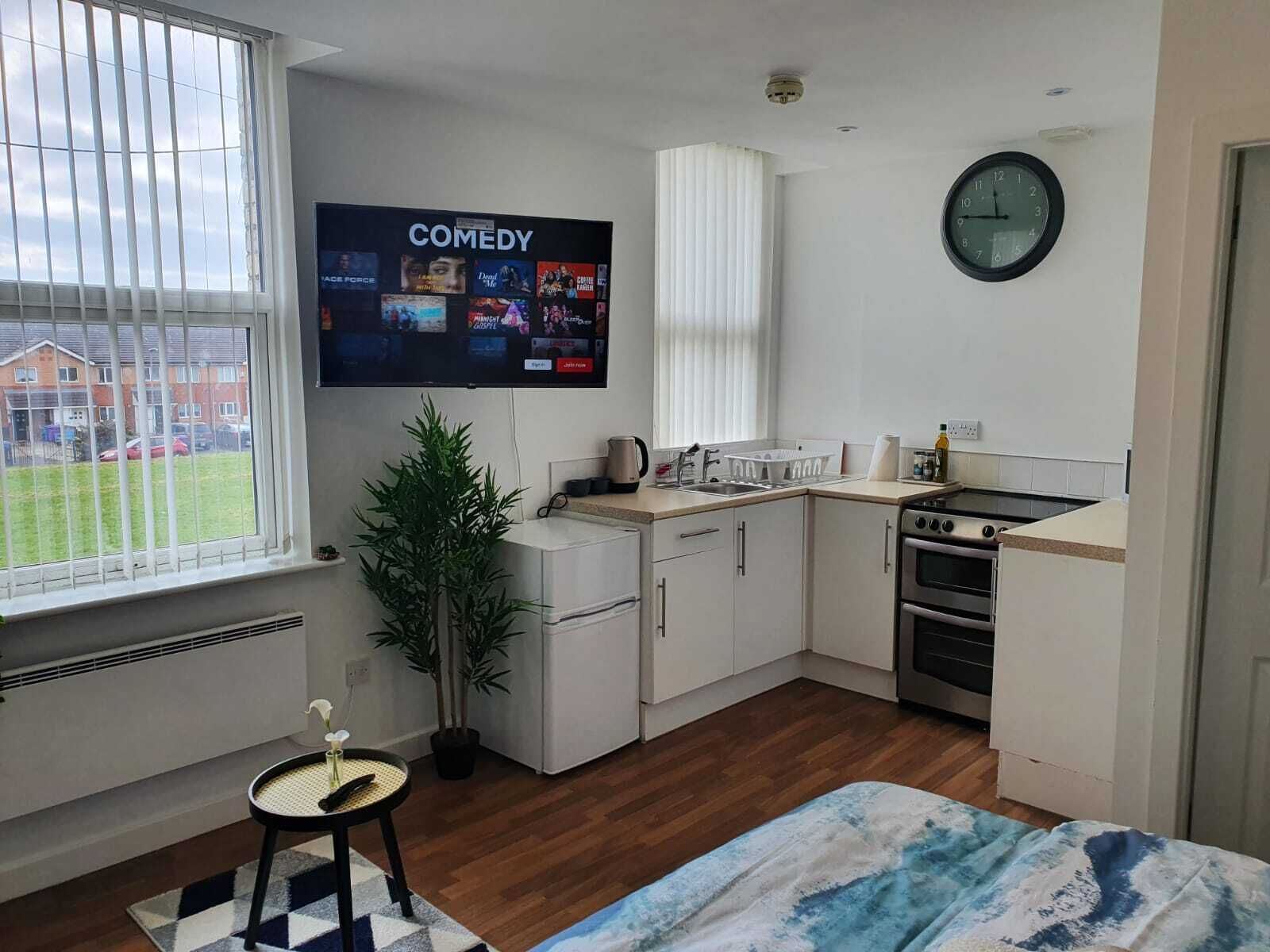 A compact kitchen with a TV displaying a comedy channel, a clock, and a view of a residential area.
