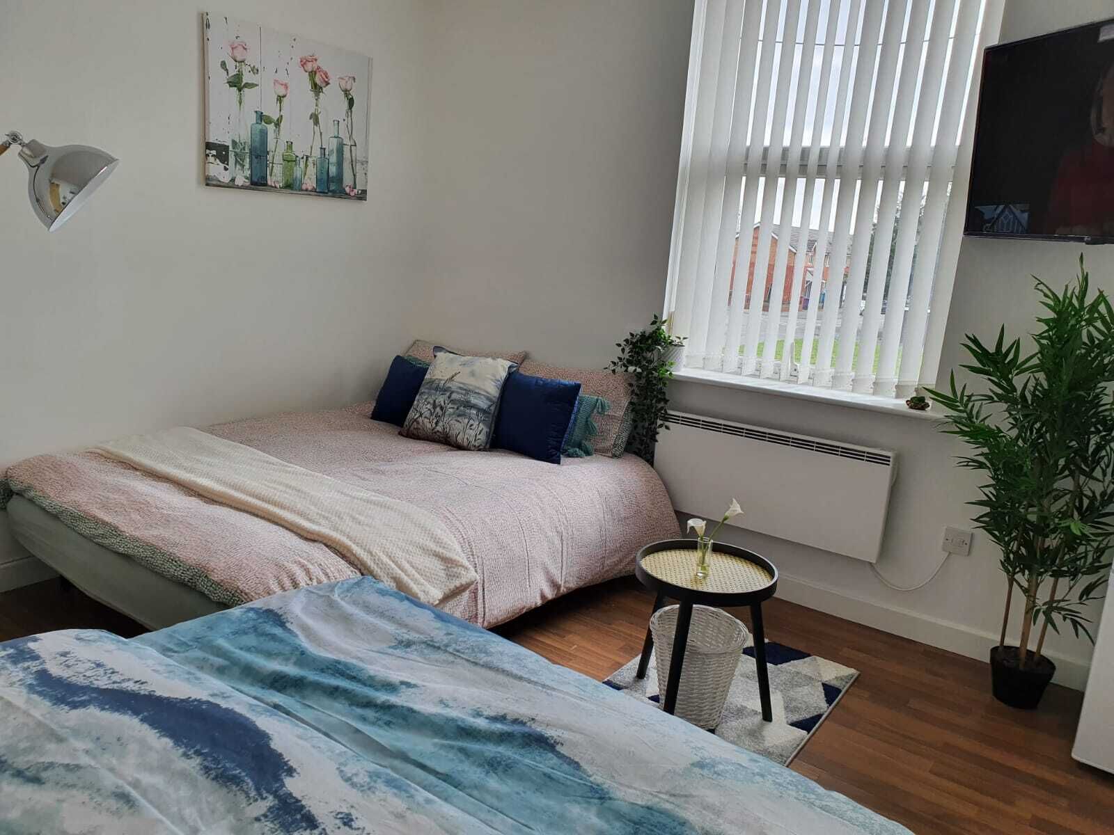 A bedroom with a bed, a small table, a potted plant, and a window with blinds.