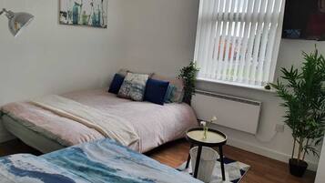 A bedroom with a bed, a small table, a potted plant, and a window with blinds.