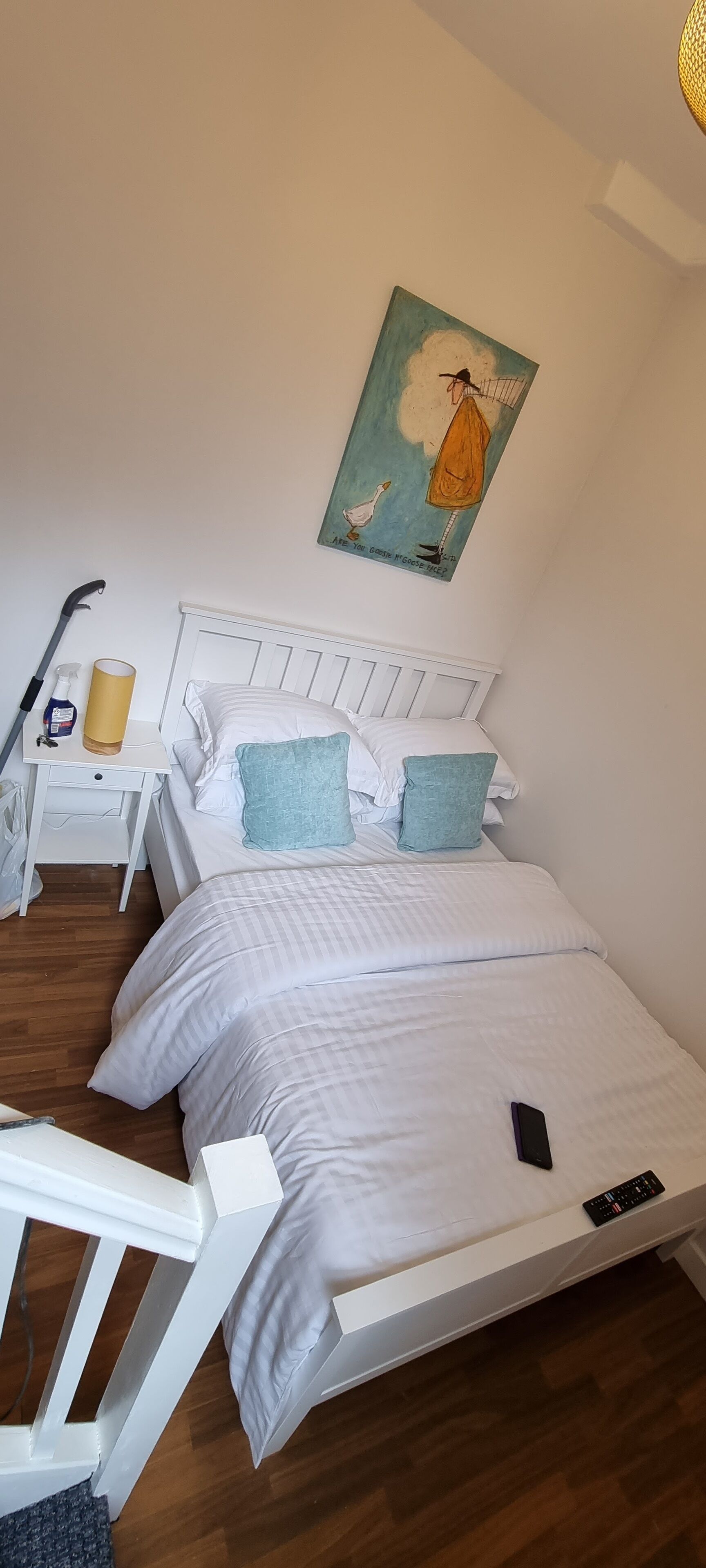 Une chambre avec un lit blanc, deux oreillers, un tableau au mur, une table de chevet avec une lampe et une télécommande sur le lit.
