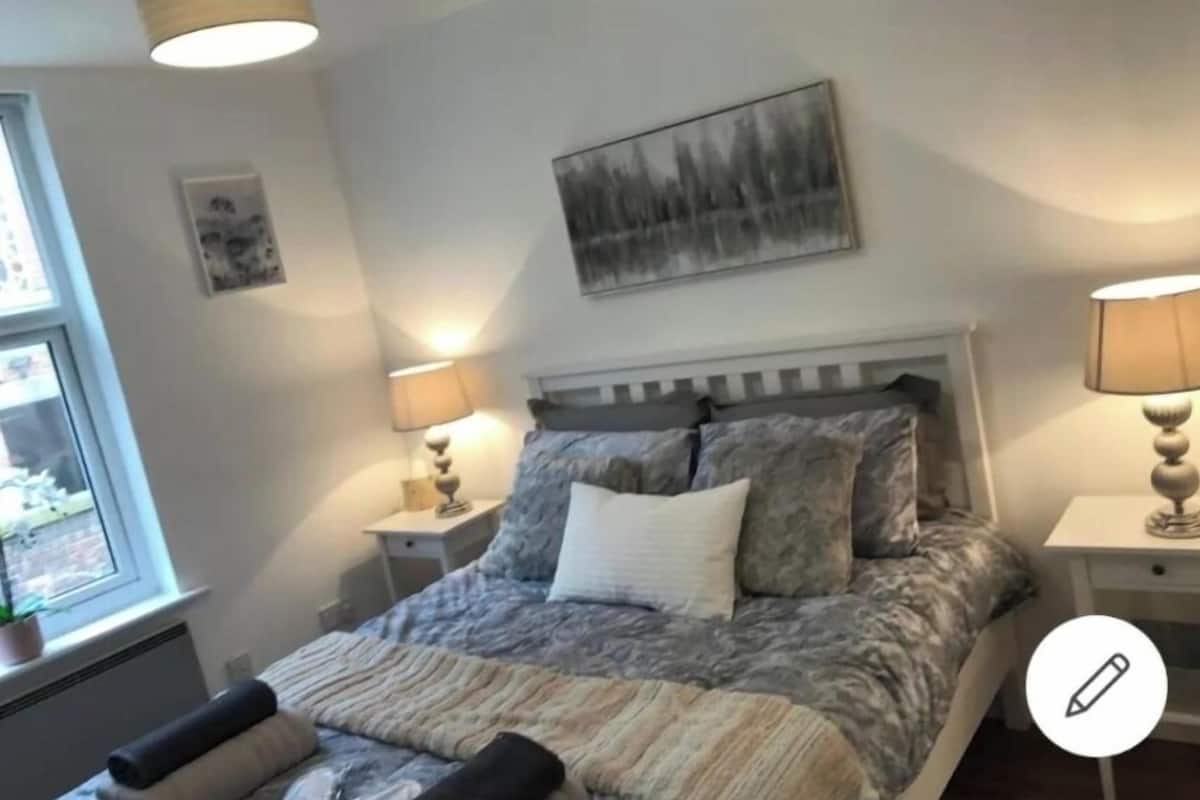 A bedroom with a bed, two bedside tables with lamps, a window, and a wall-mounted painting.