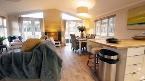 A modern living room with a sofa, a dining area with a table and chairs, a kitchen area with a counter and cabinets, and a large window with curtains. - Hollicarrs - The Rushes (York)