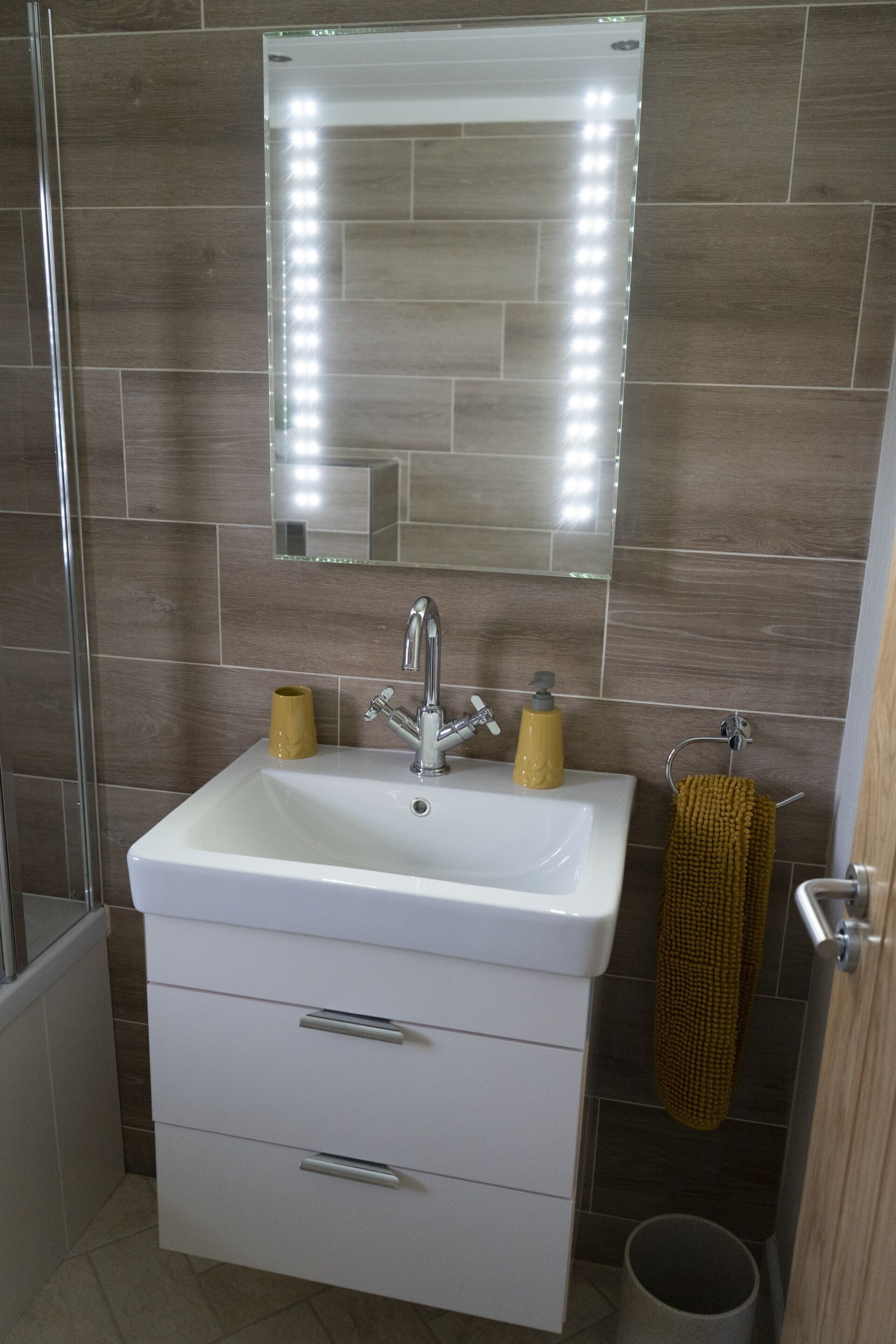 A bathroom with a white sink, a mirror with lights, a towel rack, and a trash bin.