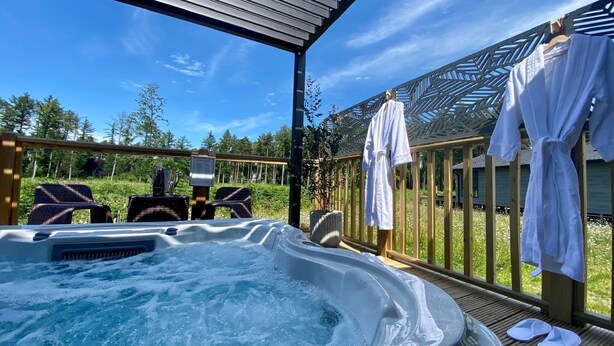 A hot tub with a wooden deck, outdoor seating, and a view of a forest. - Hollicarrs - The Rushes (York)
