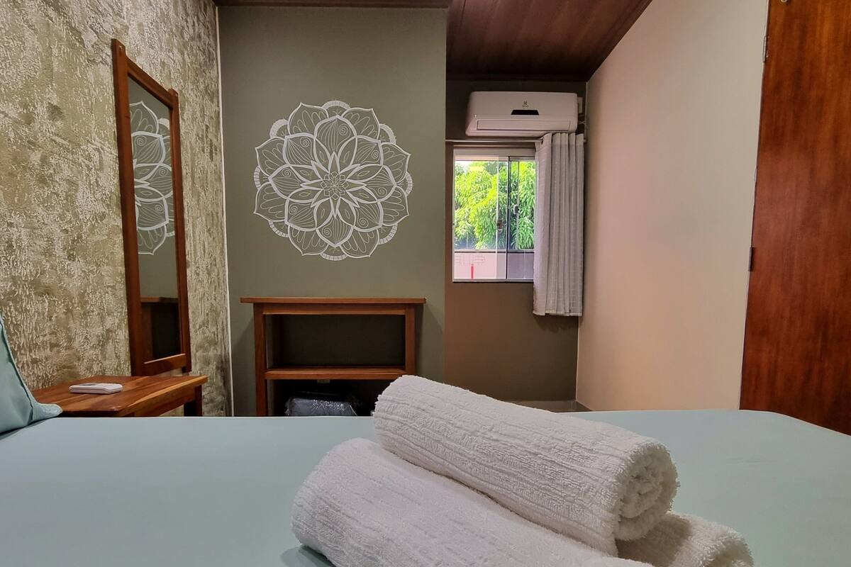 A bedroom with a bed, a wooden headboard, a wall with a decorative mandala, a window with curtains, and an air conditioning unit.