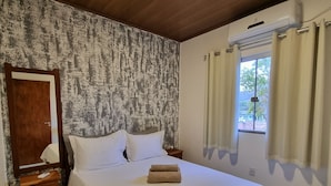 Una cama bien hecha con ropa blanca, un cabecero de madera, una mesita de noche con una lámpara y una ventana con cortinas.