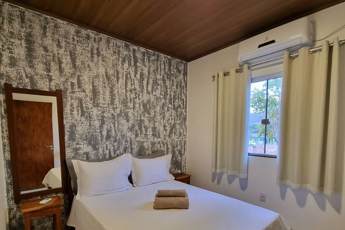 A neatly made bed with white linens, a wooden headboard, a bedside table with a lamp, and a window with curtains.